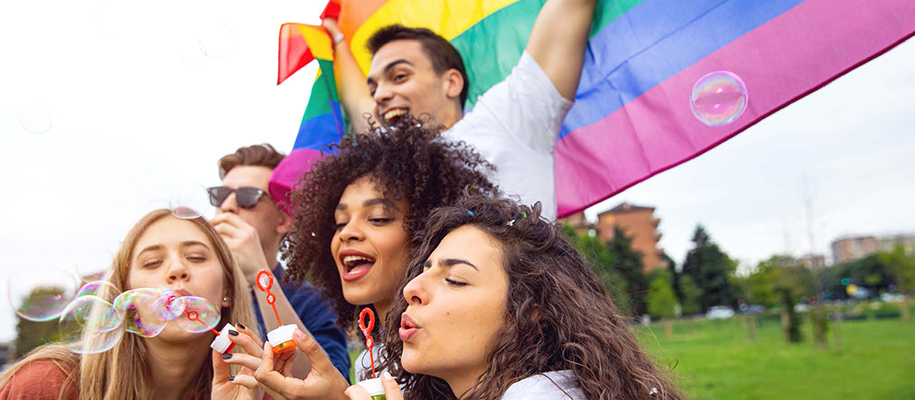 Group of diverse LGBTQ+ youth having fun, blowing bubbles, holding Pride flag