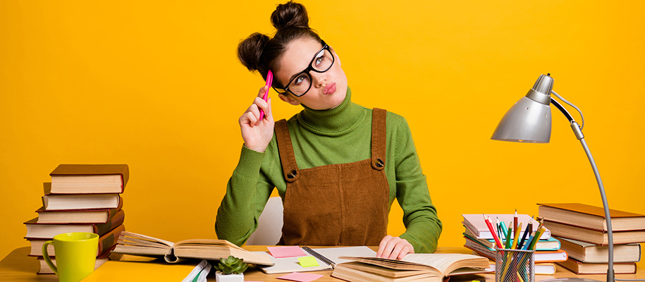 White teen in brown overalls, green shirt, hair buns studying at messy desk