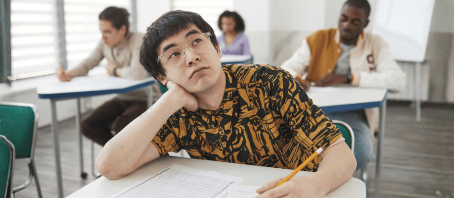 Male student sits at desk in class, taking a test, looking up contemplatively