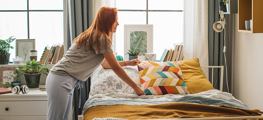Young redheaded White woman making bed with colorful pillow in sunlit dorm room