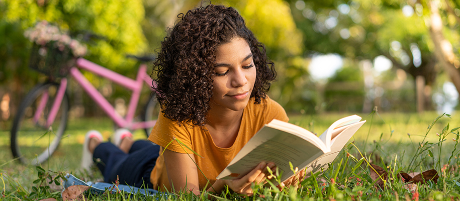 Young Black woman with curl hair in yellow shirt reading in park near pink bike