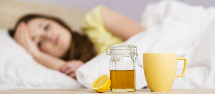 Focus on jar of honey, lemon, mug, with sick woman in bed in blurred background