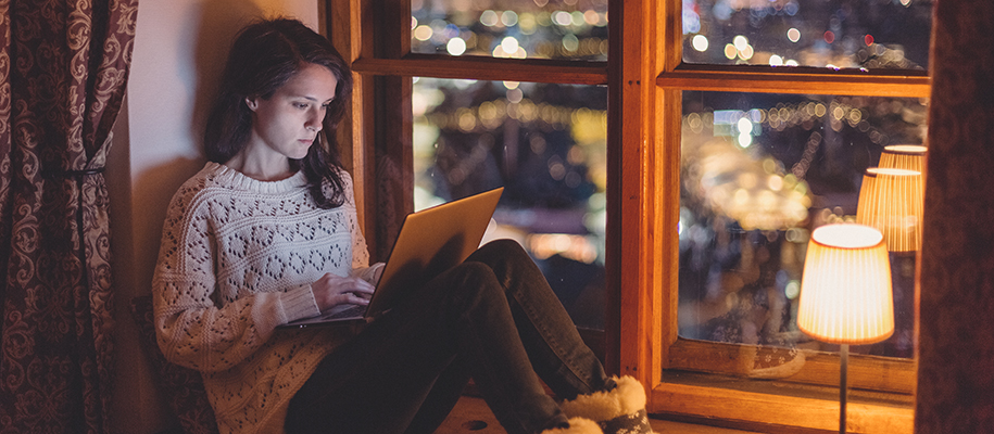 White woman in white sweater on laptop by lamp on windowsill overlooking city