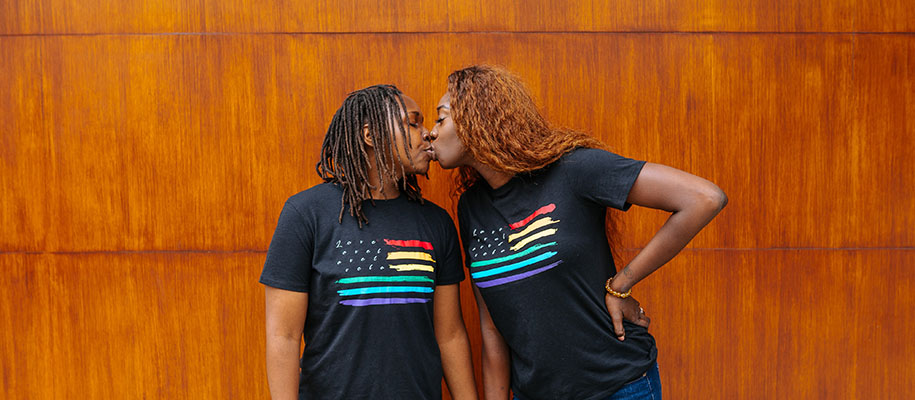 Two Black women in rainbow American flag shirts kissing in front of orange wall