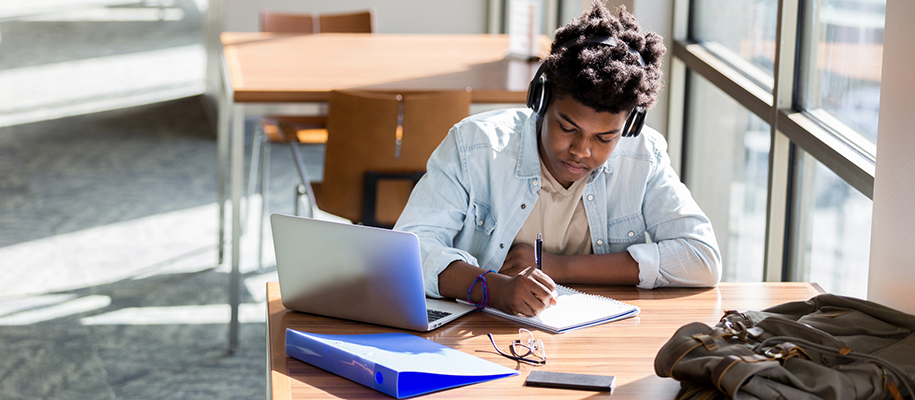 Black student wearing headphones, writing in notebook at table by large window