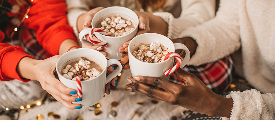 Hands of three diverse women holding festive hot chocolate mugs at pajama party