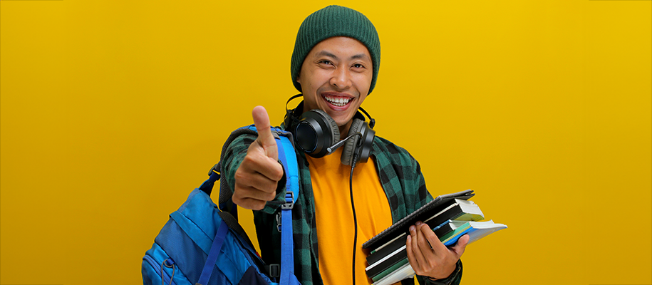 Indonesian male teen in beanie has books, bag, headphones, gives thumbs up