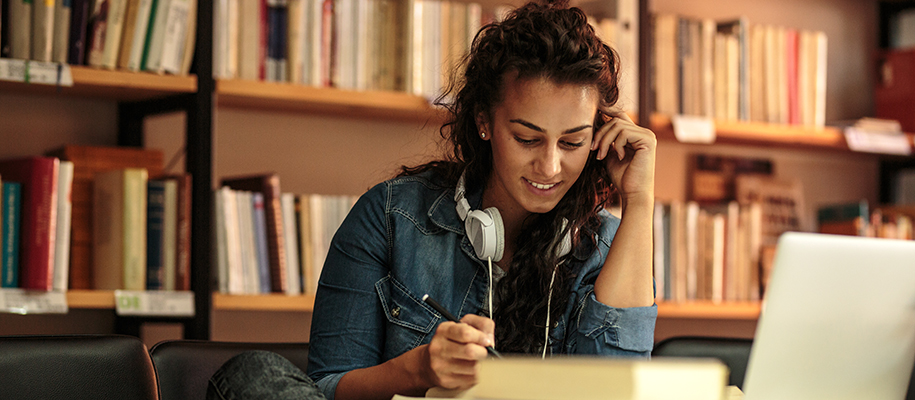 Dark-skinned female smiling to herself in library with headphones, books, laptop