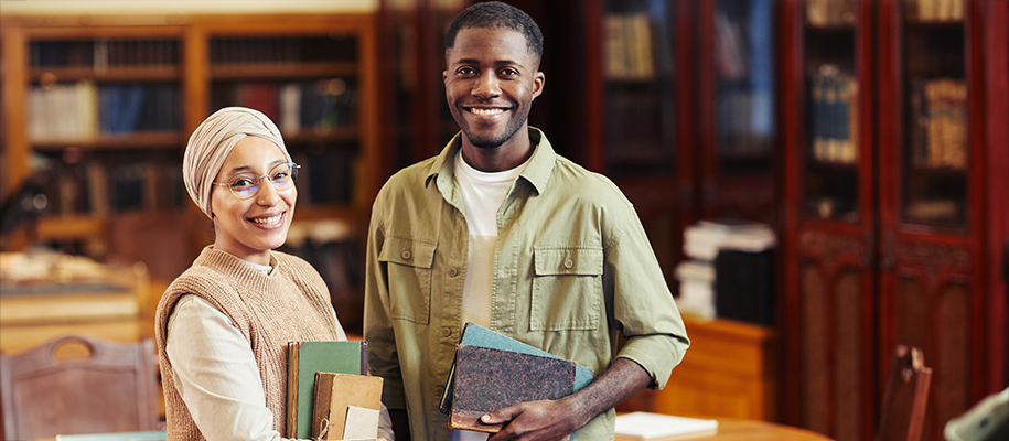 A diverse young man and woman carrying books and smiling in a library