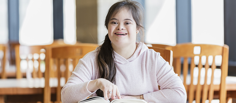 Young brunette woman with Down Syndrome in pink sweatshirt with book, smiling