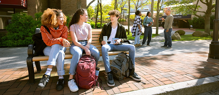 Three diverse students on campus bench talking, three people chat in background