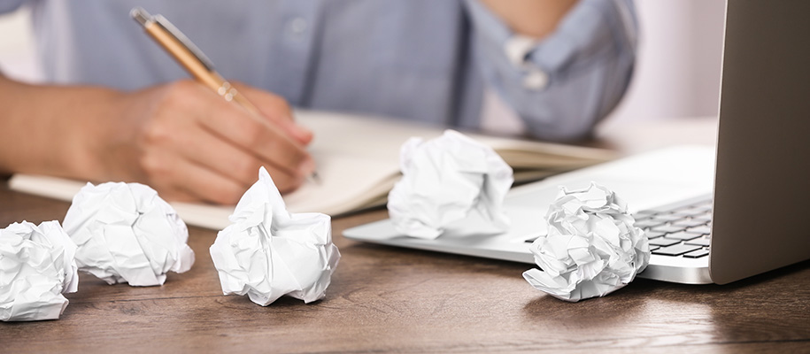 White hands write in notebook next to Macbook and 5 crumpled pieces of paper