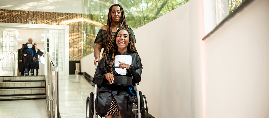 Young Black woman with braids smiling in grad gown in wheelchair pushed by mom