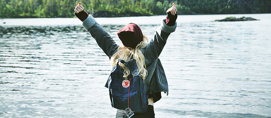 Girl in hat and fall coat with backpack raising arms in triumph in front of lake