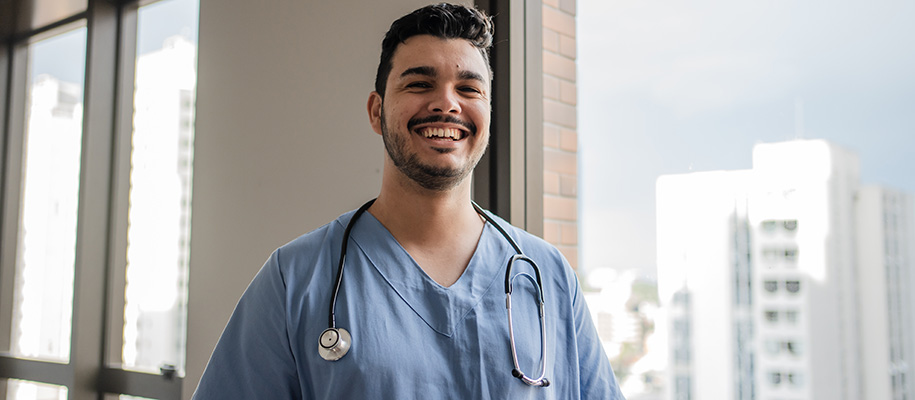 Male nurse in blue scrubs smiling and standing by window on high floor
