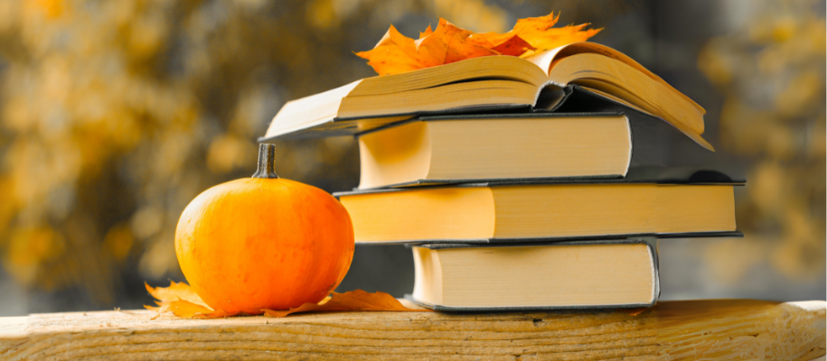 Stack of books on fence outside, top one open, with pumpkin and leaves