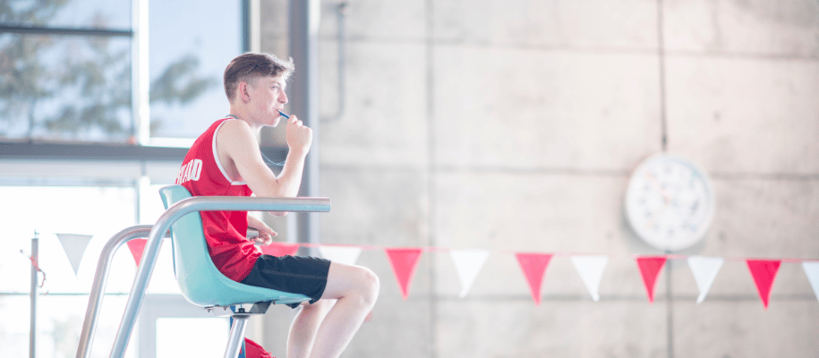 Male teen lifeguard sits on chair at indoor pool