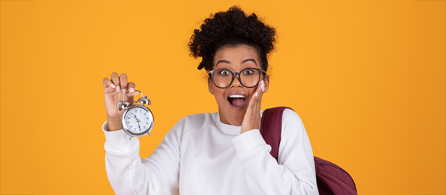 Black teen in glasses, white shirt with hand to her face, holding up alarm clock