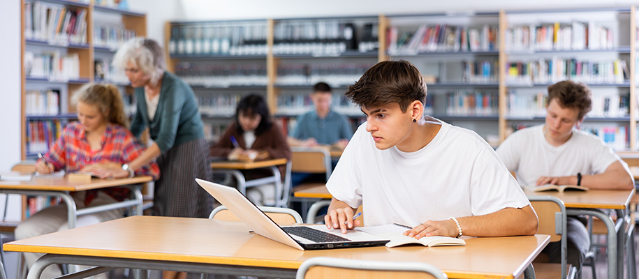 White male in white T-shirt working in library with teacher and other students