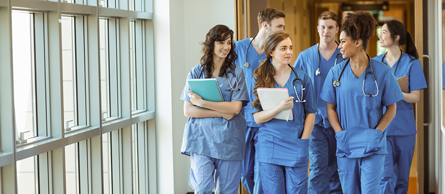 Group of nurses in blue scrubs with stethoscopes, a few with notebooks, in hall
