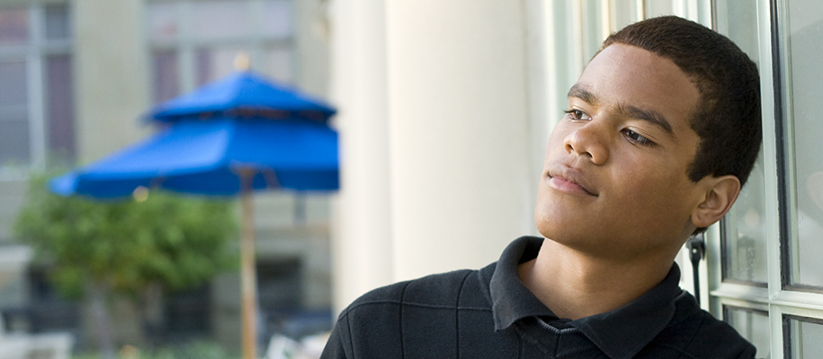 Young Black man in collared shirt leaning on window outside, looking pensive