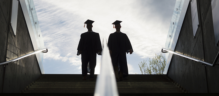 Two college graduates in caps an gowns silhouetted at top of outdoor stairs