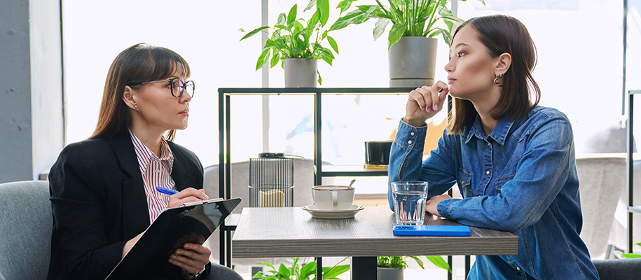 White woman in blazer and glasses with clipboard interviewing young Asian woman