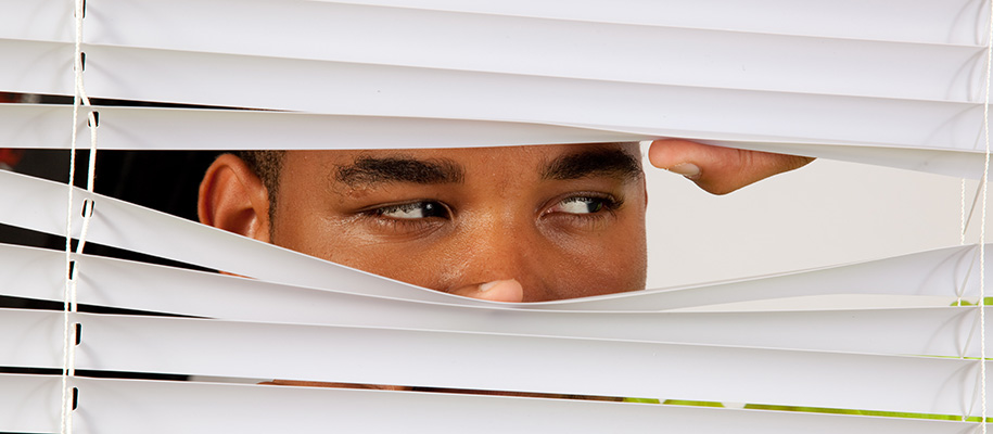 Black man peeking through white blinds looking to side