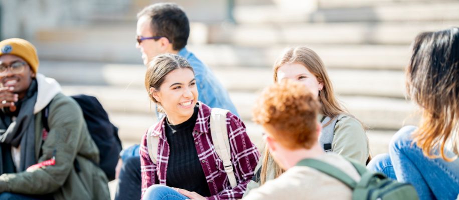 Young Latina woman in plaid shit smiling with group of students on building step