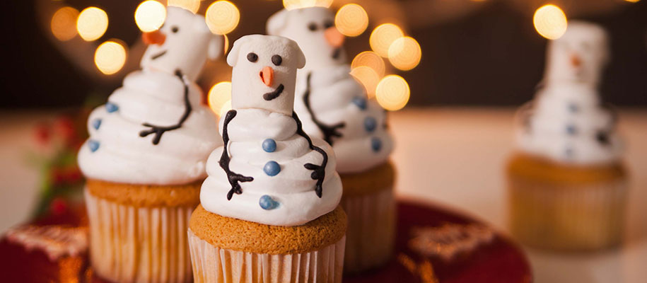 White cupcakes with snowmen made of frosting on top with lights in background