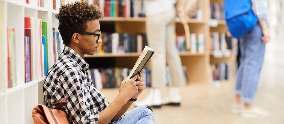 Young Black man wearing glasses and plaid shirt, reading book on library floor