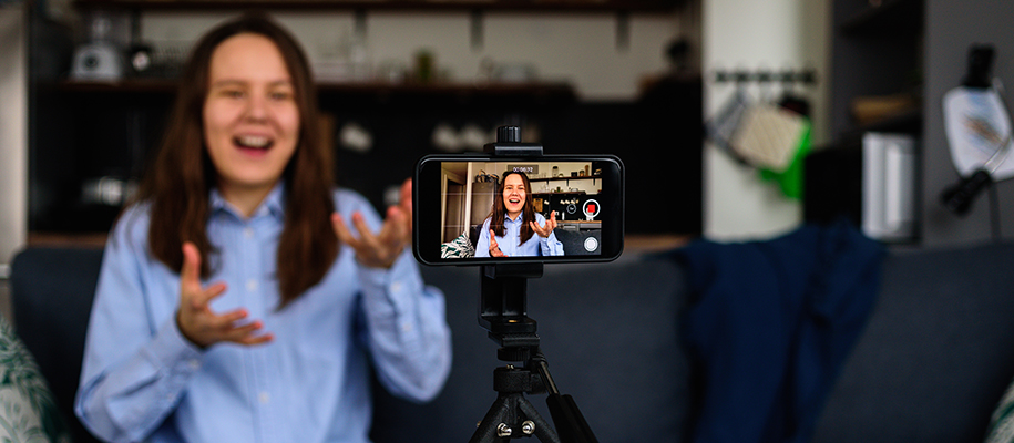 Teen with long brown hair in blue button-up shirt recording phone video on couch