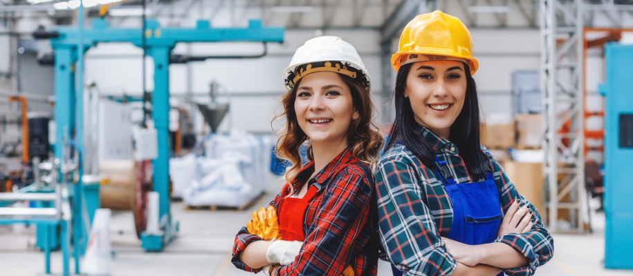 Two white woman in hard hats, plaid shirts, aprons, back to back, crossed arms