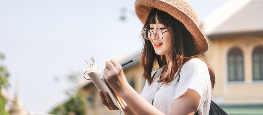 Asian woman with sun hat and round glasses, outside, writing in journal